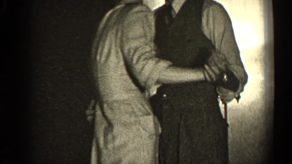 An adult couple continues dancing closely in a dim private interior, with the man's vest and the woman's dress visible.