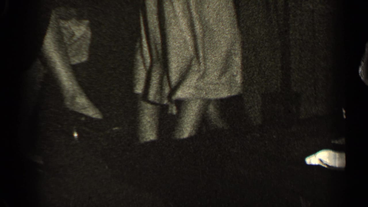 Dim indoor view of people standing near hanging coats or garments.