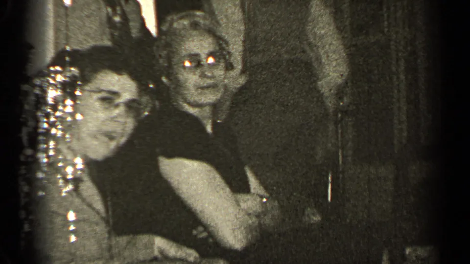 A woman with glasses smiles toward the camera in a 1947 Illinois indoor gathering, with another guest and reflective decorations nearby.