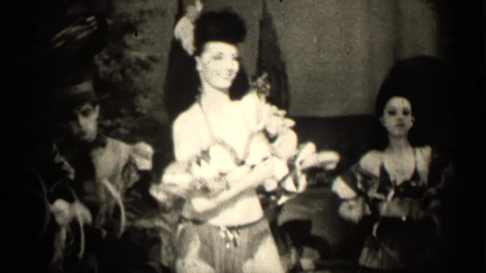 A woman in a feathered hat appears to perform indoors in 1947 Illinois, with other costumed or dressed-up women visible around her.