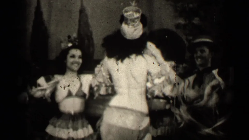 In 1947 Illinois home-movie footage, a woman in a grass skirt smiles beside a clown-costumed performer and another costumed adult.