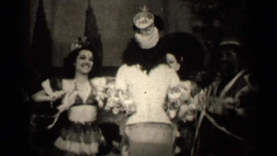 A spectator-shot 8mm frame shows the clown-costumed performer from behind, with the grass-skirted woman and another performer partly visible.