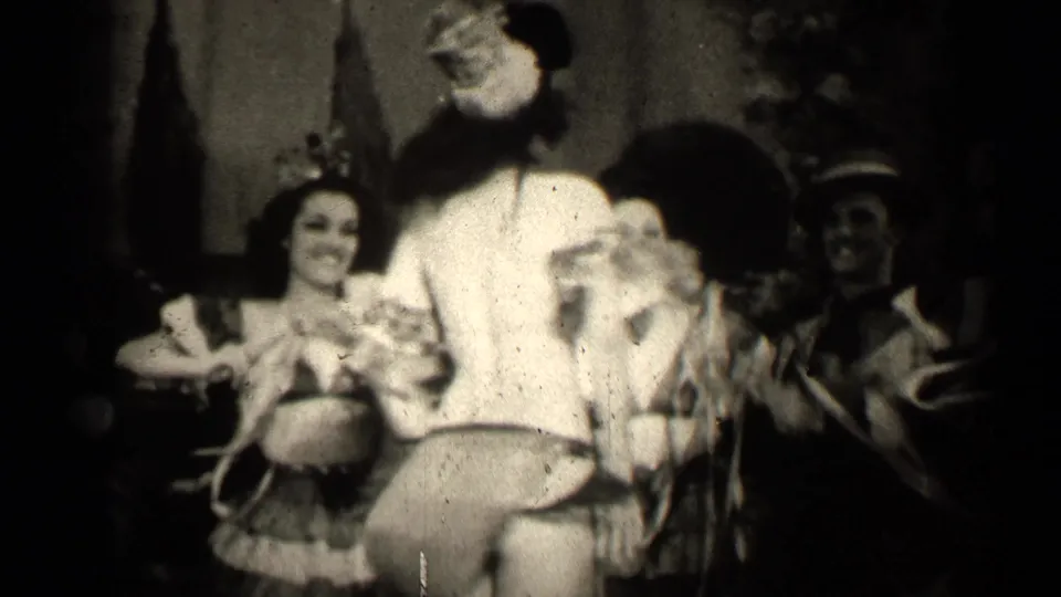 In the final frame, the clown-costumed performer turns partly toward camera while the grass-skirted woman smiles beside other costumed adults.