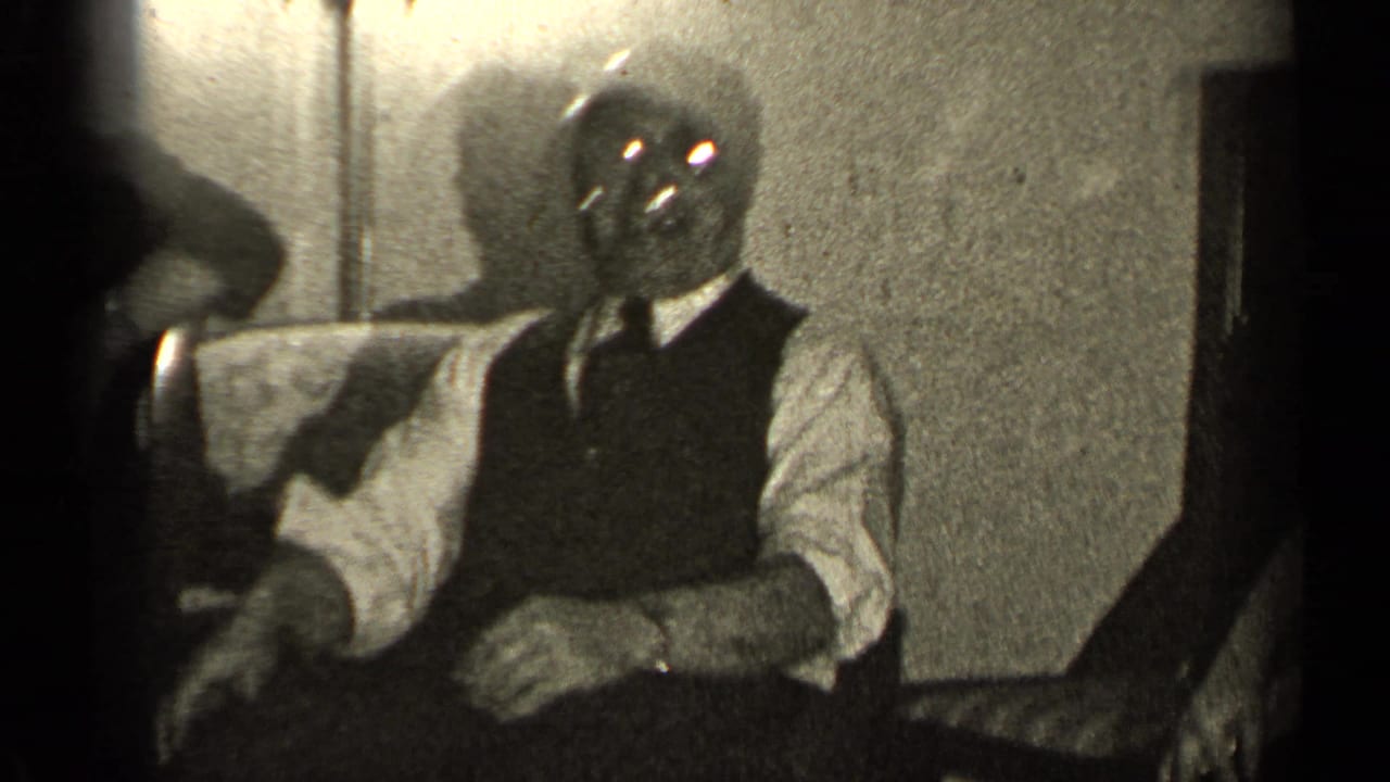 In the Illinois home-movie setting, a seated man in a vest and tie faces the camera in a dim private room.