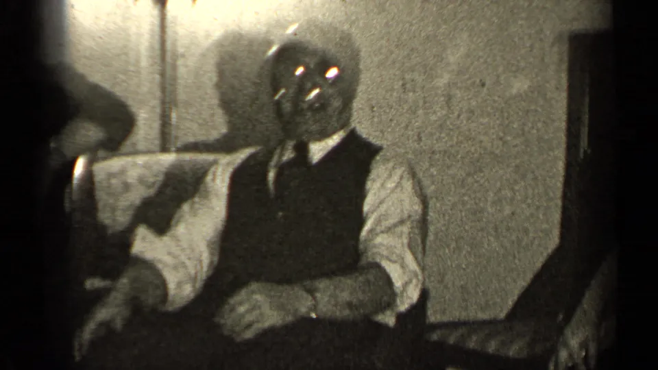 In the Illinois home-movie setting, a seated man in a vest and tie faces the camera in a dim private room.