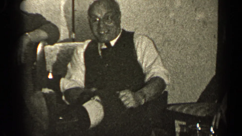 A man in a vest and tie sits in a private Illinois interior, with another person partly visible at the edge of the frame.