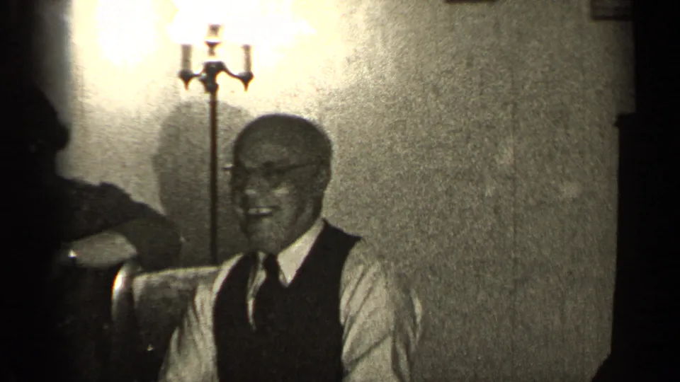 A smiling man in a vest and tie sits near a floor lamp in the 1947 Illinois home-movie interior.