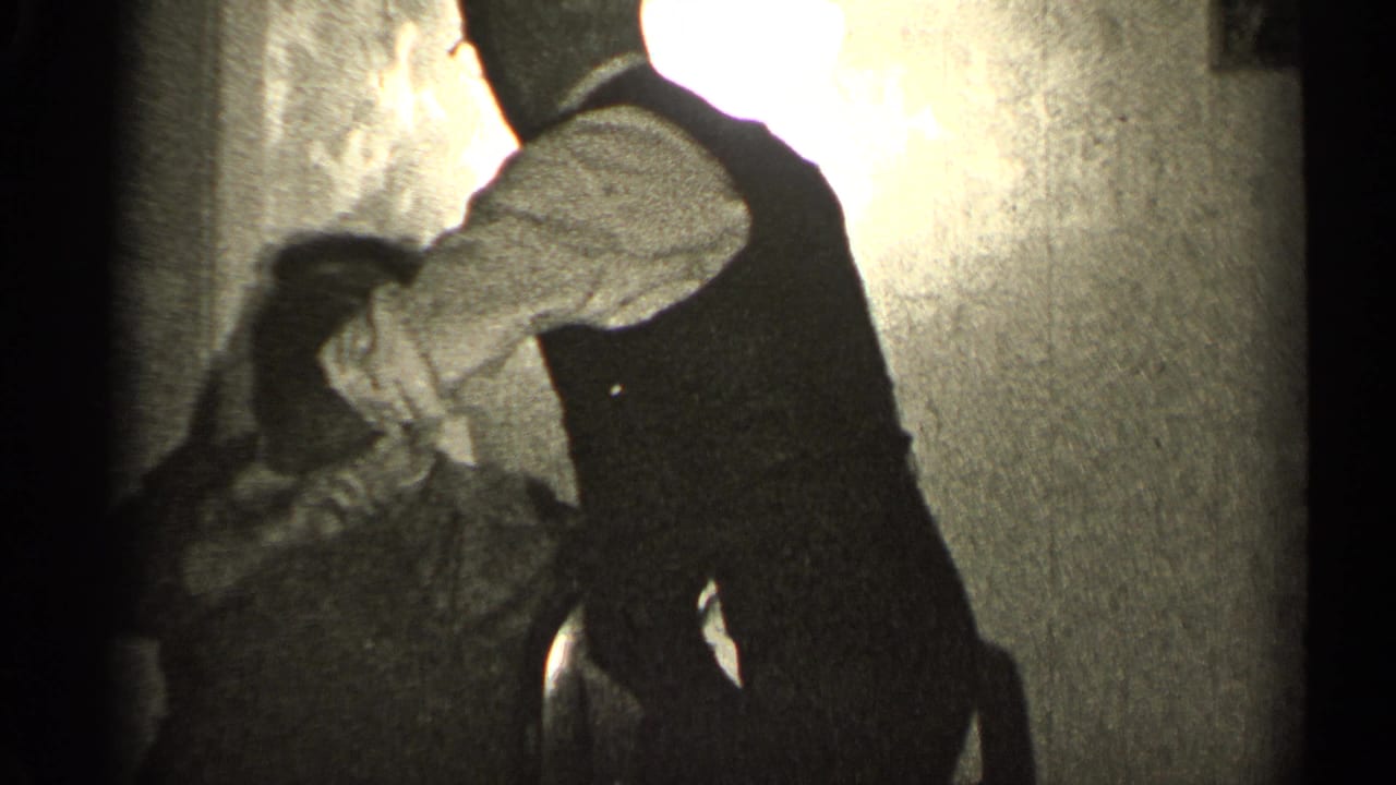 Man in a dark vest leans over a seated woman in a dim interior.