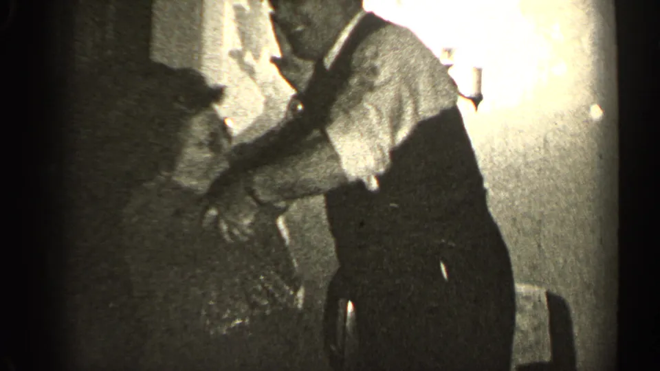 In the Illinois home interior, the man bends toward the seated woman, with a wall lamp and chair visible behind them.