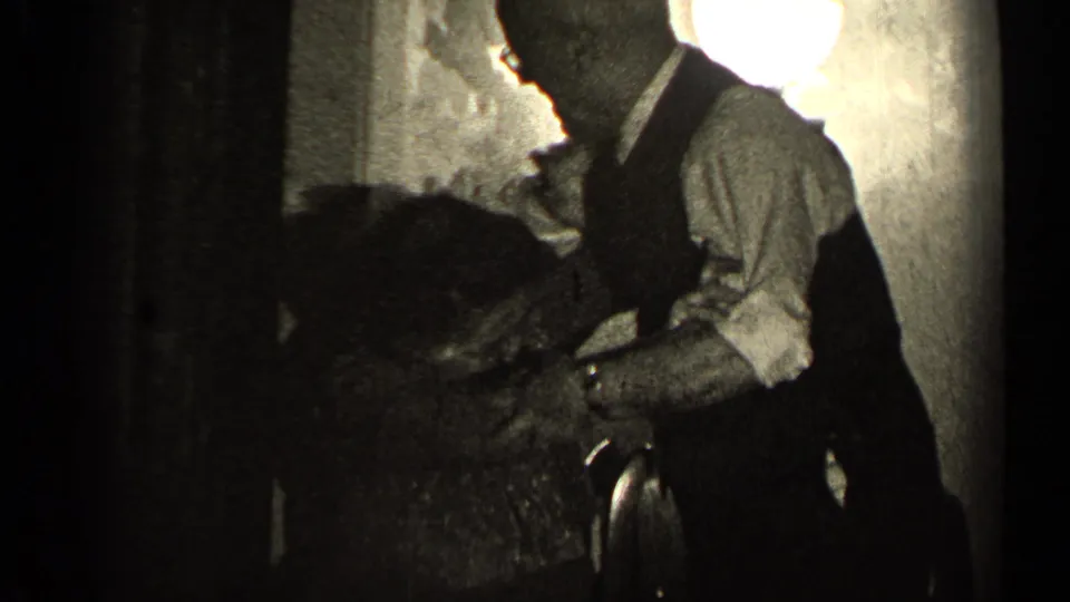 In 1947 Illinois home-movie footage, an older man in a vest bends toward an older woman in a low-lit private room.