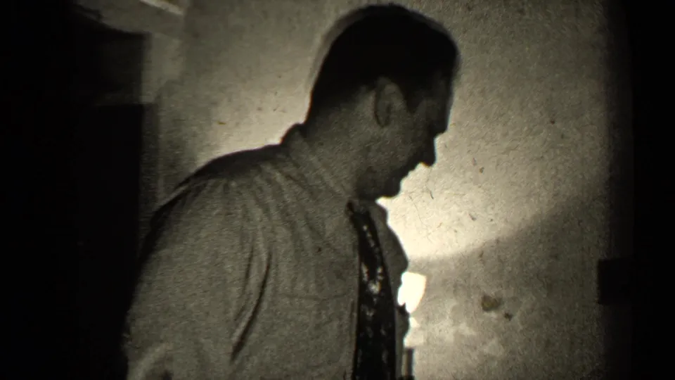 In 1947 Illinois home-movie footage, a man in a collared shirt and tie stands in profile near a lamp in a dark interior.