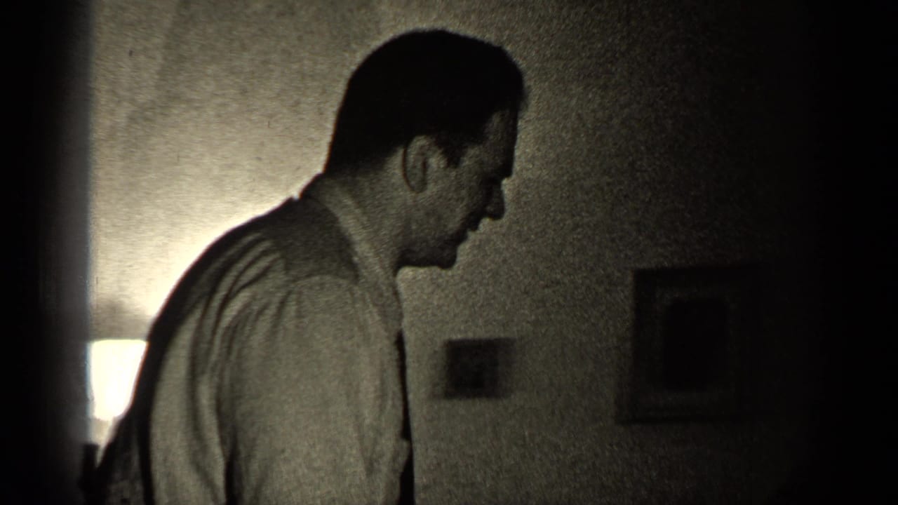 The man remains in side profile in a dim Illinois interior, with a glowing lamp and small framed wall pictures behind him.