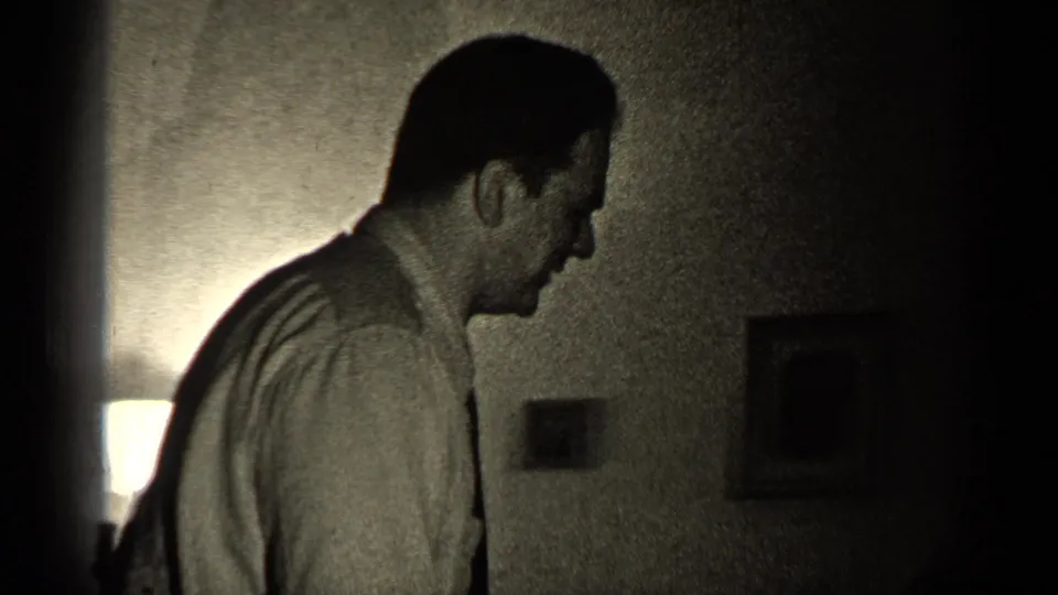 The man remains in side profile in a dim Illinois interior, with a glowing lamp and small framed wall pictures behind him.