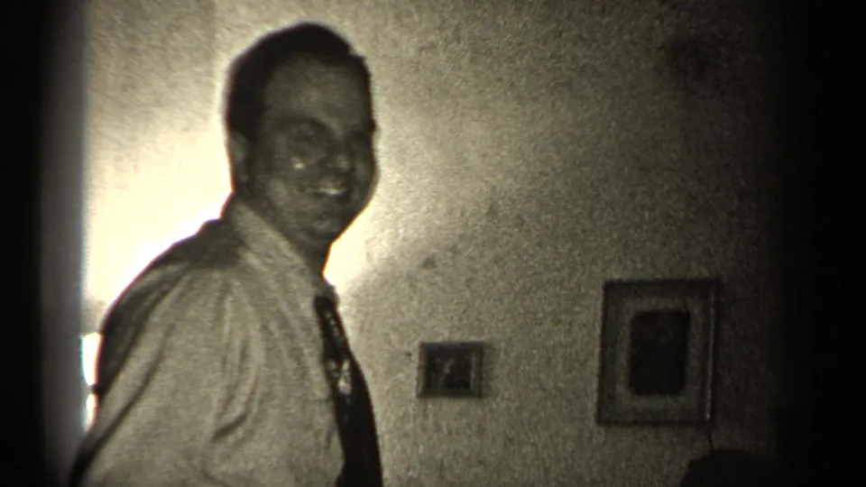 The man turns toward the camera with a smile, wearing a shirt and loosened tie in a softly lit 1947 Illinois interior.