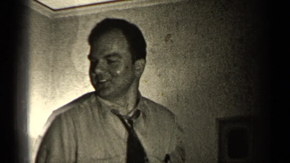 A man in a shirt and loosened tie looks off to the side in a dim 1947 Illinois home interior with framed wall decor.