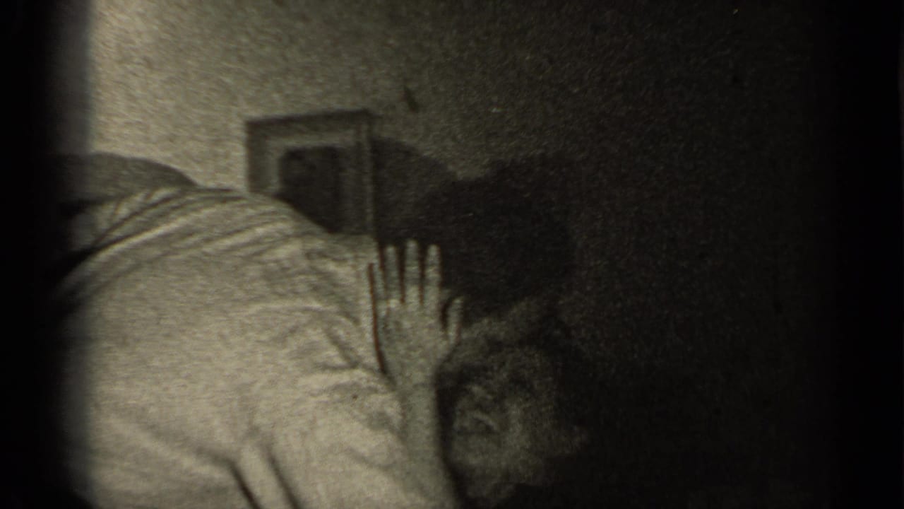 Man leans over a woman lying on a bed in a dark room.