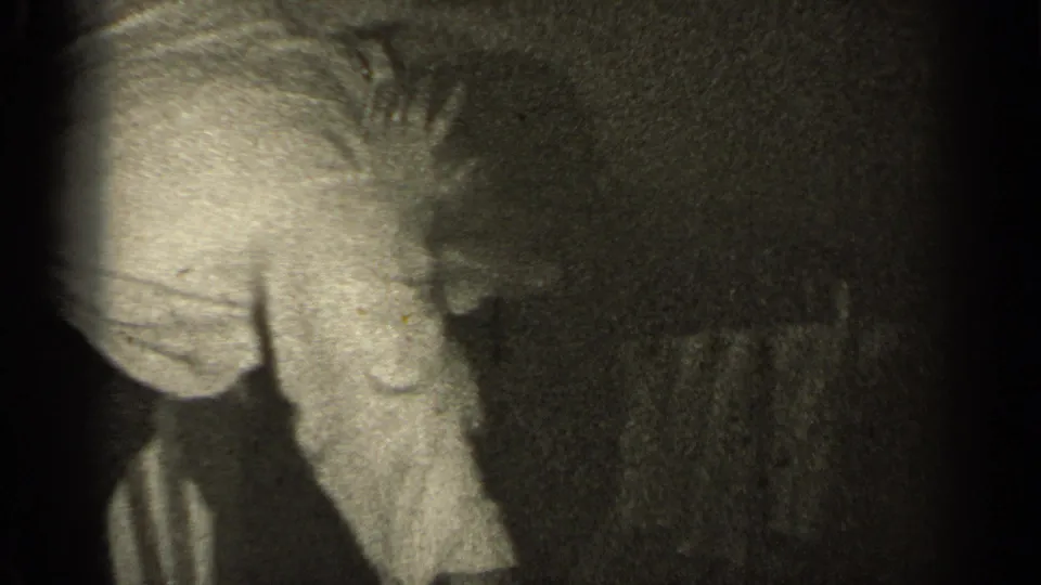 A close, grainy home-movie frame shows the man bending over the reclining woman in a dark bedroom-like interior.