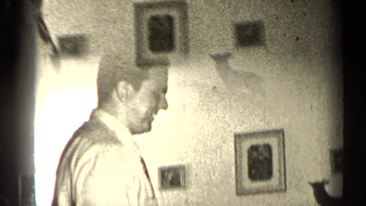 Man in a light shirt stands in a dim room with framed pictures on the wall.