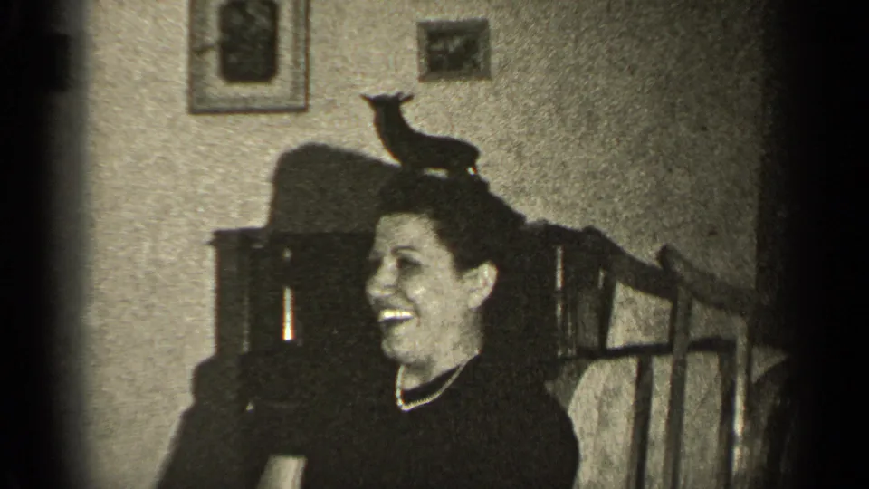 A smiling woman sits in a 1947 Illinois interior, with framed pictures, dark furniture, and a small animal-shaped object nearby.