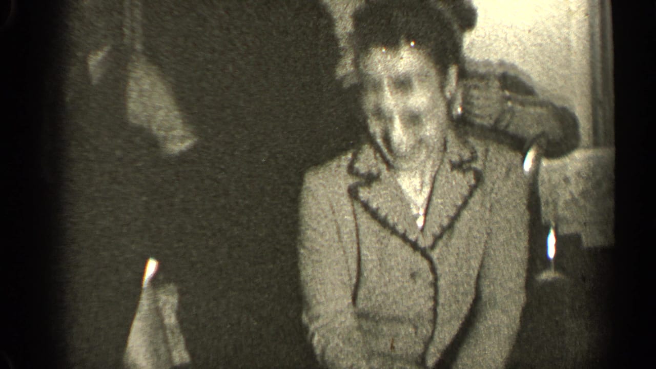 Woman in glasses and a light coat stands in a dim room holding a white handkerchief.