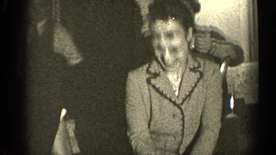 In a 1947 Illinois home-movie frame, a woman in glasses and a light coat stands indoors with a white handkerchief nearby.