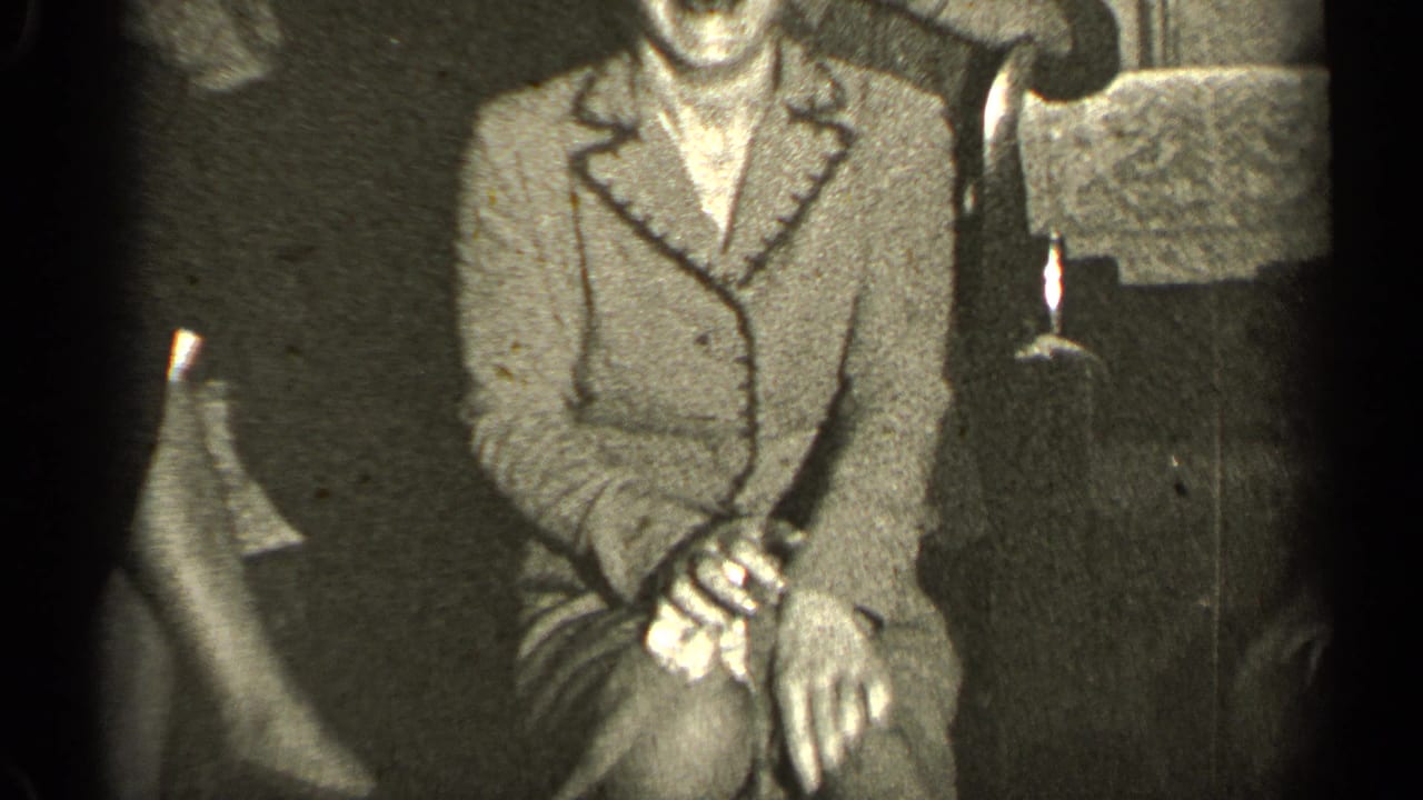 Woman in a light coat stands with her hands together beside an upholstered chair.