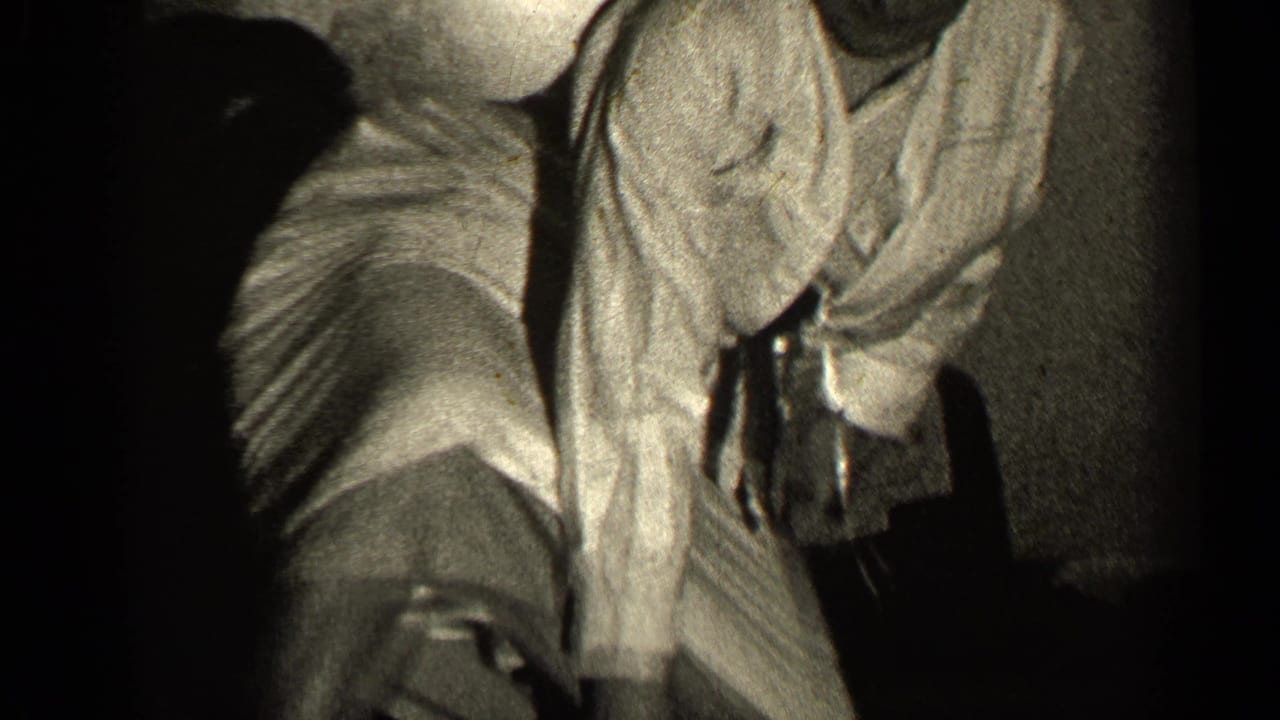 Woman in a light suit crouches low near a chair in a dark interior.