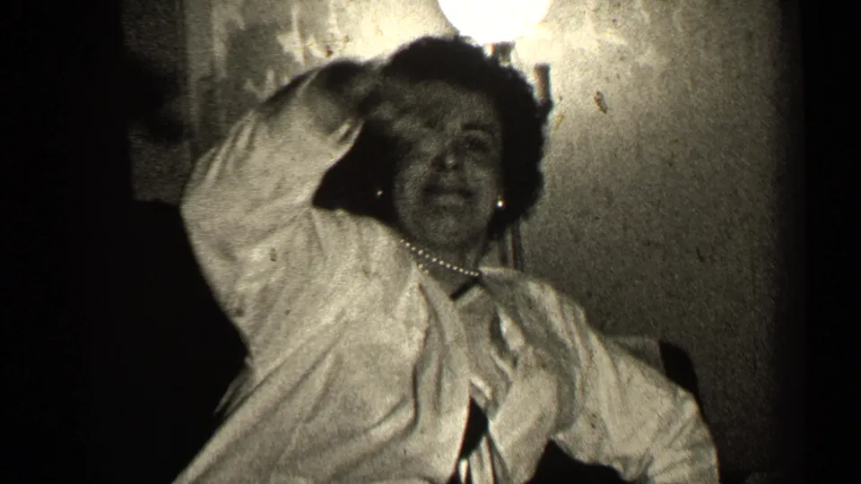 The woman raises a hand to her hair or head while seated or leaning near the chair in the 1947 Illinois interior.