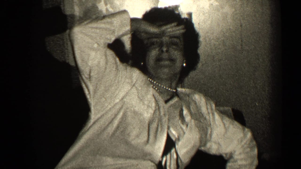 Woman in a light dress and pearl necklace raises one arm while seated indoors.
