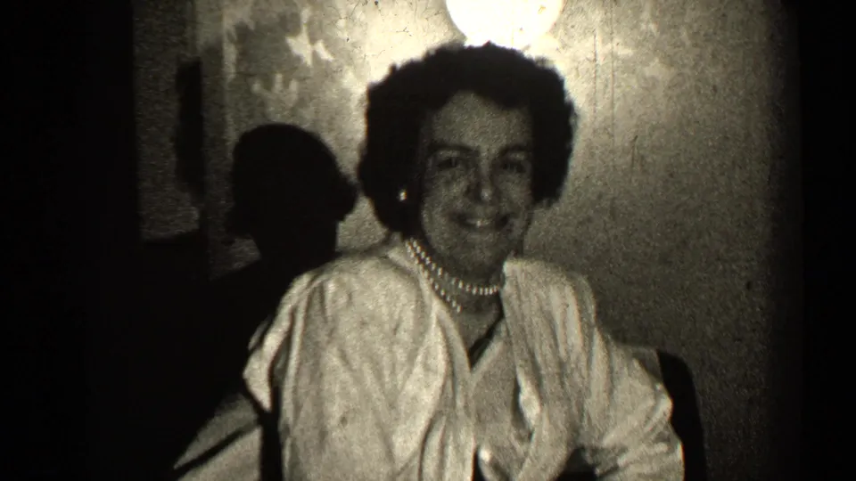 In 1947 Illinois home-movie footage, the woman smiles broadly toward the camera in a dim private interior.