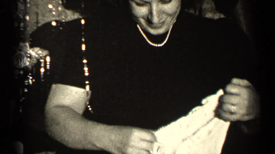 In 1947 Illinois home-movie footage, a woman wearing pearls looks down while holding light-colored fabric indoors.