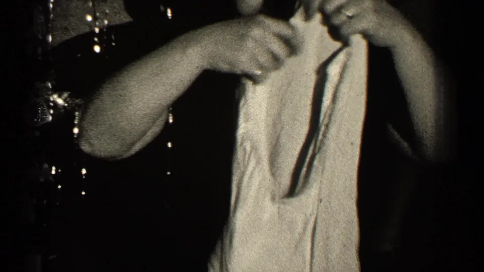 In the black-and-white home movie frame, the woman raises the light-colored garment as if checking its shape or fit.