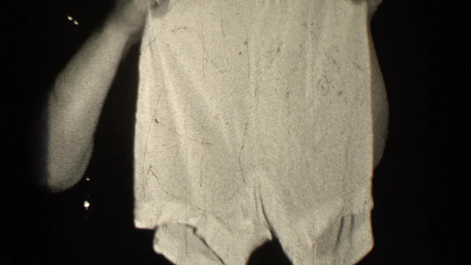The home-movie frame shows a close view of a light textured garment held up in a dim 1947 Illinois interior.
