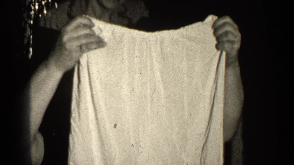 The woman remains mostly obscured by the light shorts as she holds the garment up in the dark, high-contrast 8mm frame.