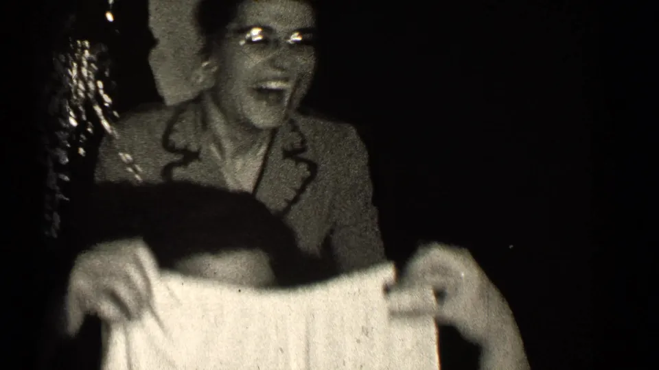 A woman smiles or laughs behind the raised shorts, her face visible in the high-contrast 1947 Illinois home-movie frame.