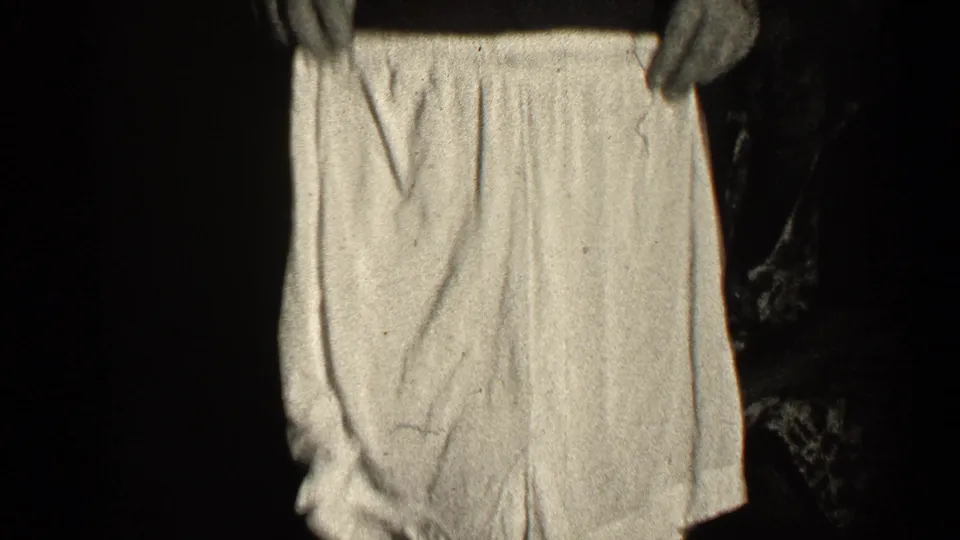 A person holds a light-colored undergarment up to the camera in the dim black-and-white home-movie frame.