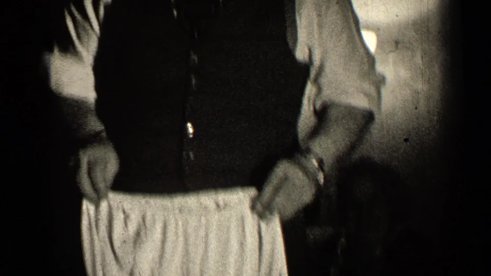 Inside a dim Illinois home-movie scene, cropped figures stand close as a light undergarment is held at waist height.