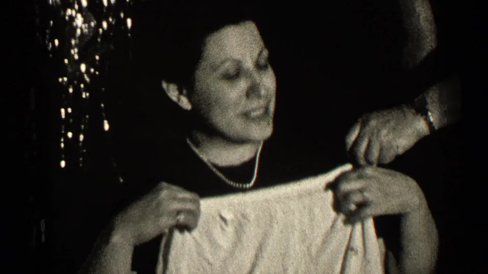 A middle-aged woman in a dark dress and necklace holds a light garment as another adult hand gestures nearby.