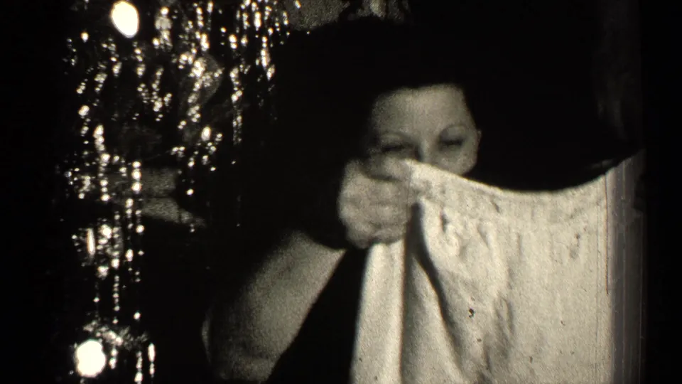 The woman raises the pale garment near her face in dim 1947 Illinois home-movie footage, with reflective lights at left.