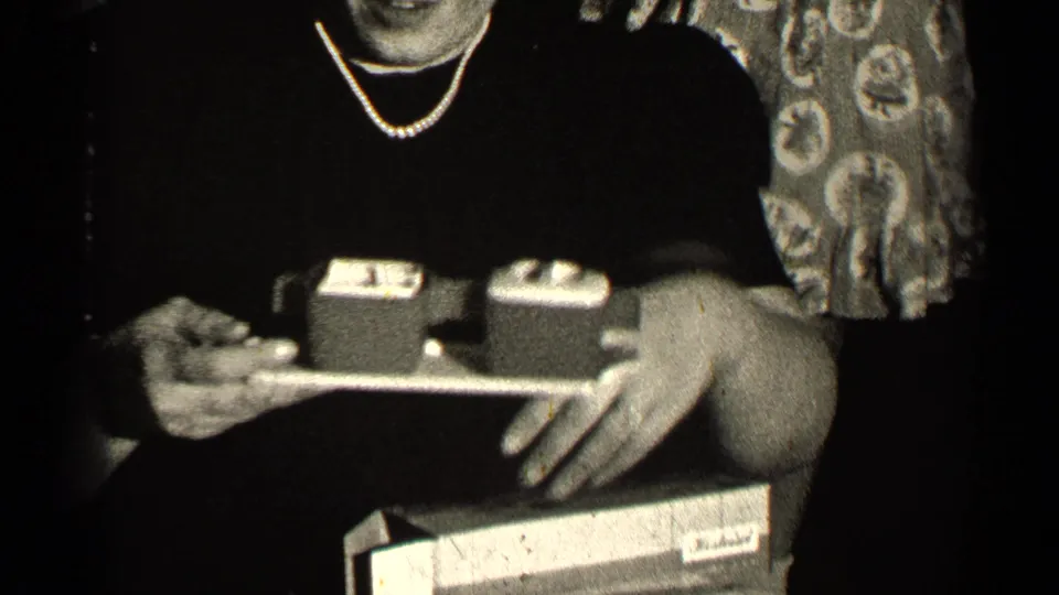 A woman in a dark dress and pearl necklace holds a tray with small gift boxes in 1947 Illinois bridal shower footage.