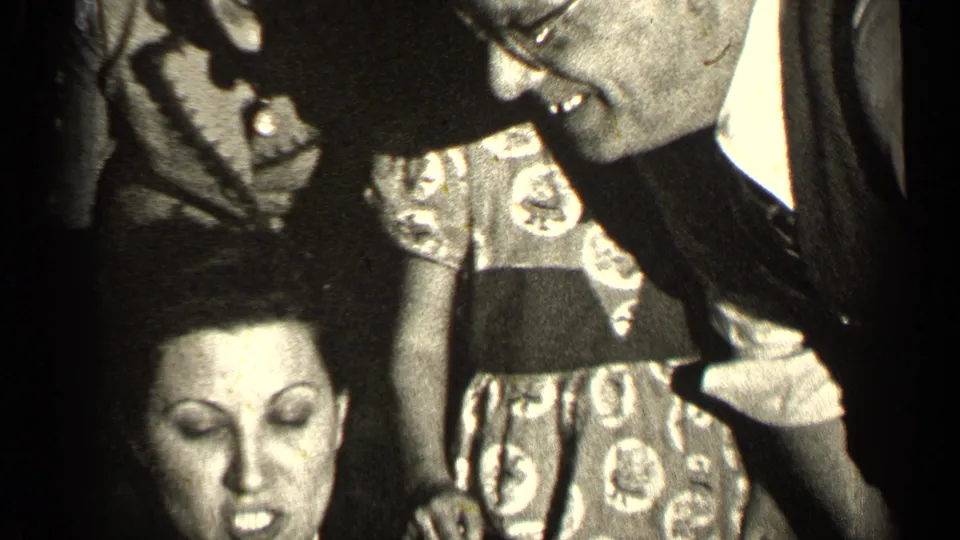 A suited man with glasses leans close during the family gathering, with the woman in pearls below and a young girl in a patterned dress nearby.