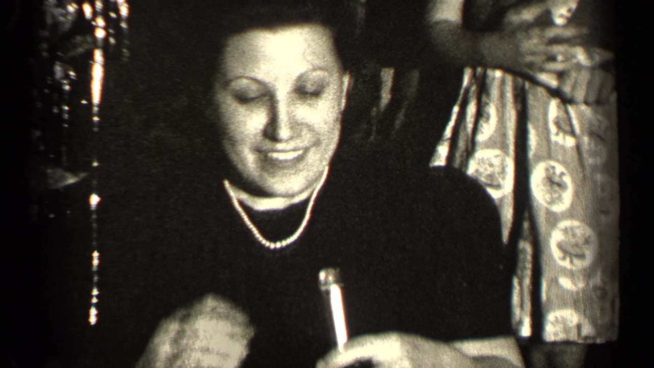 Woman in a dark dress and pearl necklace holds a small bottle at an indoor gathering.