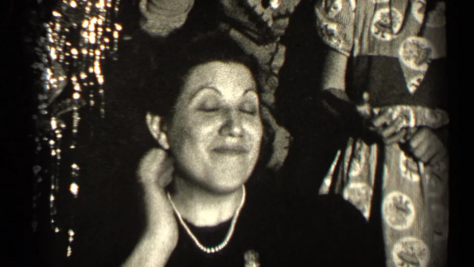 The woman closes her eyes and touches her neck, appearing to enjoy the scent during the 1947 Illinois indoor gathering.