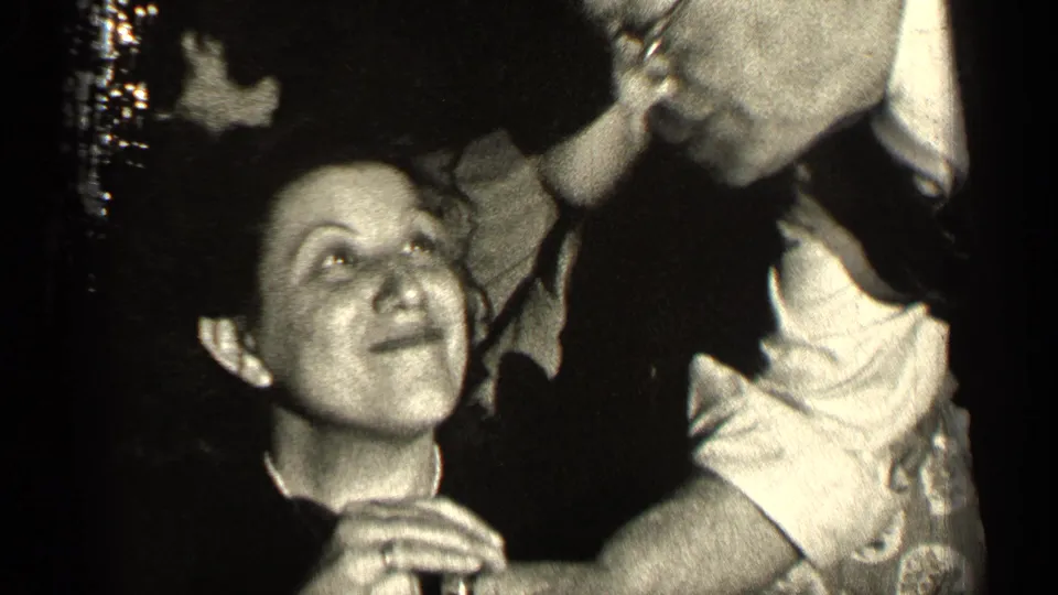 At the Illinois gathering, the seated woman looks up as a man leans close, with hands and party clothing filling the frame.