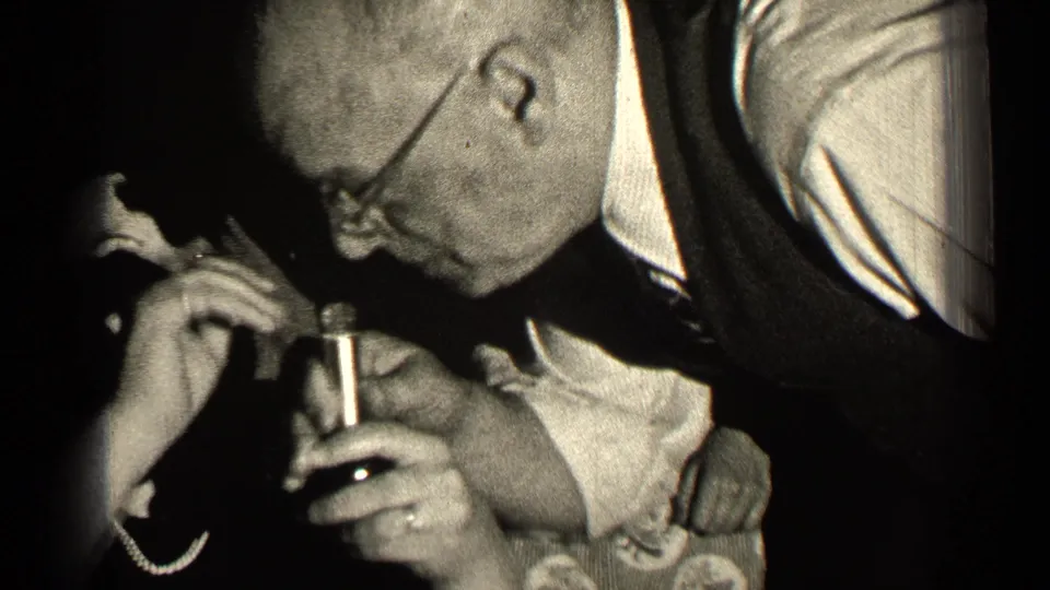 A man wearing glasses leans toward the small perfume bottle while the woman and others remain close in the 1947 Illinois home movie.