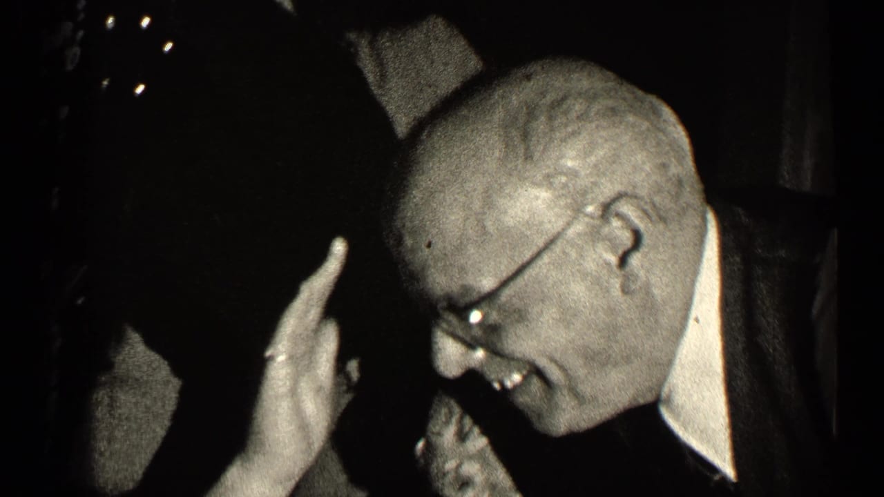 Man in glasses laughs in a dark interior while raising one hand near his head.