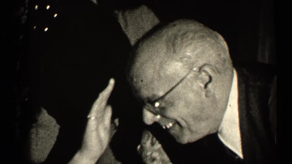 In a 1947 Illinois home-movie frame, a man in glasses and a dark suit laughs in a dim room with his hand raised.