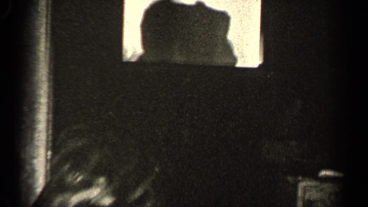 A dim black-and-white interior frame shows a bright window-like opening and partial figures or shadows, with details obscured by low light.