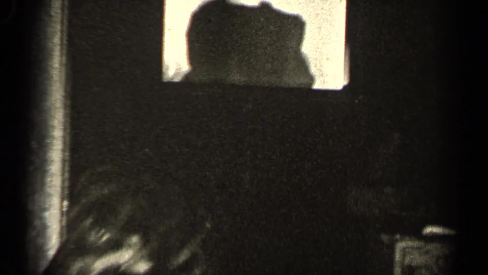A dim black-and-white interior frame shows a bright window-like opening and partial figures or shadows, with details obscured by low light.