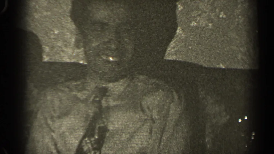 Grainy 8mm frame from 1947 Illinois with a suited man and a woman seated together on a couch in a shadowed interior.